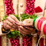 Engagement Ring ceremony- Indian Hindu male putting ring on bride’s decorated finger. Couple is well attired as per traditional Indian Hindu wedding. Groom wearing Jodhpuri suit and floral garland.